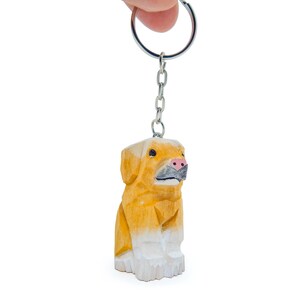 Peut inclure: Un porte-clés en bois en forme de chien golden retriever assis. Le chien a un nez rose et des pattes blanches.