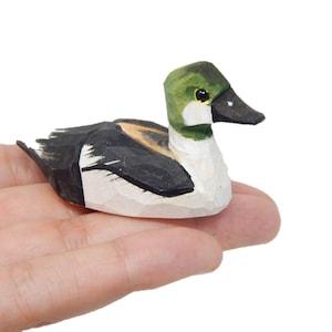May include: Hand-carved wooden figurine of a goldeneye duck. The duck is painted with black, white, and green details. The duck is sitting on a hand.