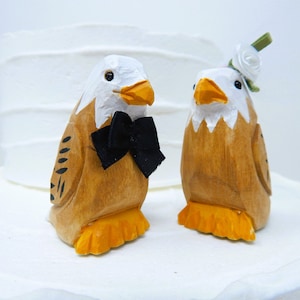 May include: A pair of wooden bald eagle wedding cake toppers. The groom eagle wears a black bow tie and the bride eagle has a white flower on her head.