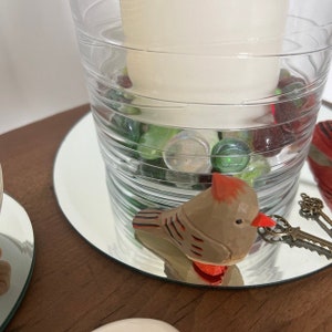 A wooden bird figurine with red accents sits on a mirrored surface. The bird is perched on top of a glass vase filled with green and red glass marbles. The vase is surrounded by a white candle.