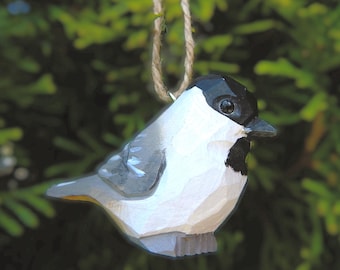 Puede incluir: Adorno de pájaro de madera tallada a mano con cabeza negra, cuerpo blanco y alas grises. El pájaro está suspendido por una cuerda de hilo natural. El fondo es verde borroso, lo que sugiere un entorno natural al aire libre. Una decoración encantadora y rústica.