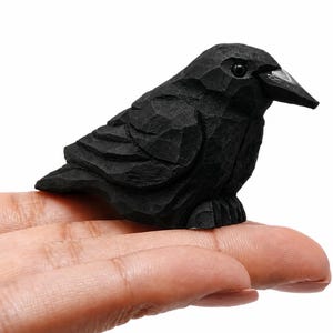 May include: A small, hand-carved wooden bird figurine finished in a matte black color. The sculpture features visible faceted chisel marks across its surface, a pointed beak, and a single dark, glossy eye, capturing the form of a perched crow.
