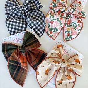 May include: Four large hair bows on white cards. The bows feature various patterns: black and white gingham, floral, plaid, and a cream-colored pumpkin design. Each bow is tied in the center and has long, flowing tails.