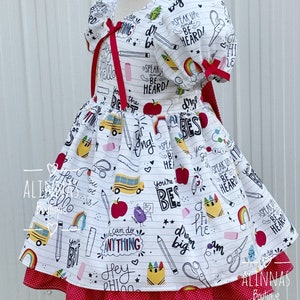 May include: White dress with a red polka dot skirt and a white bodice featuring a school-themed print with phrases like "Speak Up and Be Heard!" and "You're the Best!"