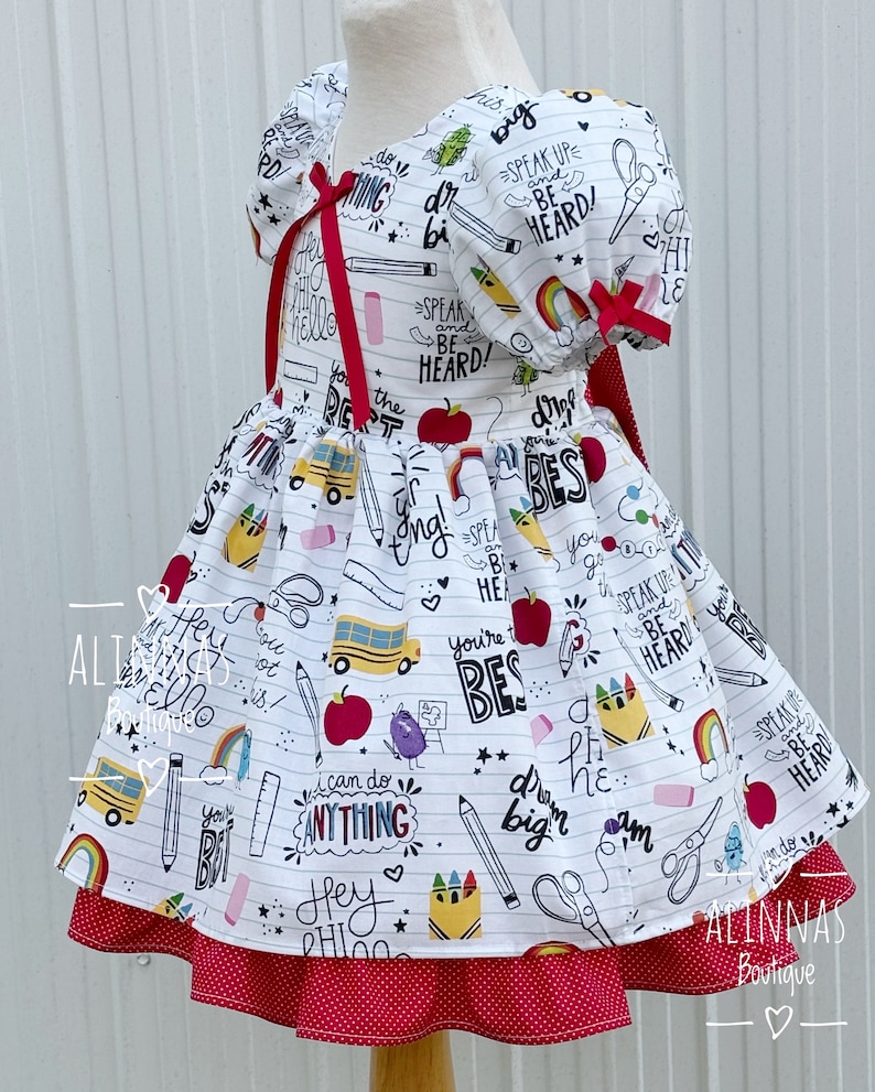 May include: White dress with a red polka dot skirt and a white bodice featuring a school-themed print with phrases like "Speak Up and Be Heard!" and "You're the Best!"