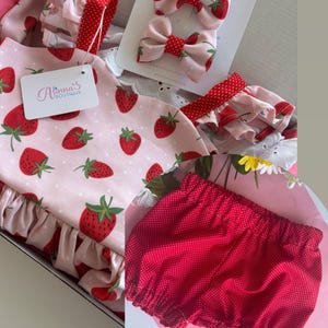 May include: A baby outfit set featuring a pink strawberry-printed dress with ruffled sleeves and a matching red polka-dot diaper cover. Includes two strawberry bow hair clips and a tag that reads "Alinna's Boutique."