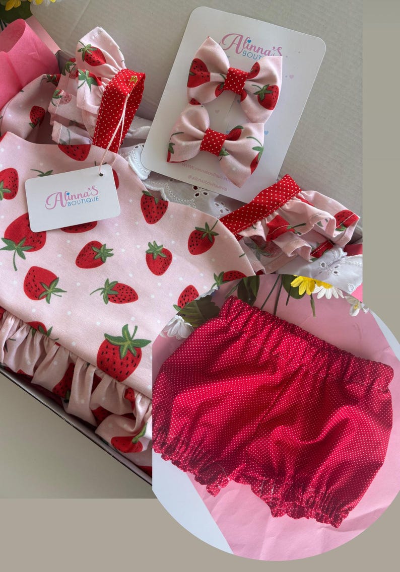 May include: A baby outfit set featuring a pink strawberry-printed dress with ruffled sleeves and a matching red polka-dot diaper cover. Includes two strawberry bow hair clips and a tag that reads "Alinna's Boutique."
