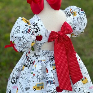 May include: White dress with a red bow and red polka dot bow. The dress has a school-themed print with words like "Hey, Hi, Hello, You're the Best, Speak Up, Be Heard, I Dream, I'm the Best, You Got This, Hard!" and images of a school bus, apple, rainbow, pencil, eraser, scissors, and a star.