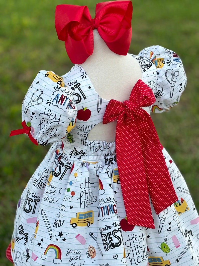 May include: White dress with a red bow and red polka dot bow. The dress has a school-themed print with words like "Hey, Hi, Hello, You're the Best, Speak Up, Be Heard, I Dream, I'm the Best, You Got This, Hard!" and images of a school bus, apple, rainbow, pencil, eraser, scissors, and a star.