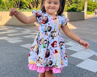 May include: A white short-sleeved dress featuring a colorful all-over print of various animated princess characters, accented with small blue and pink floral motifs. The dress has a gathered waist and a solid pink ruffled hemline at the bottom.