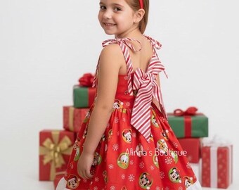 May include: A red Christmas dress with a white eyelet hem, featuring cartoon characters and snowflakes. The dress has a large striped bow in the back and a matching red headband. Gift boxes are in the background.