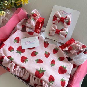 May include: A pink baby outfit set with a strawberry pattern, including a top, matching hair bows, and a ruffled trim. The set is presented in a pink box, with the brand name "Alinna's Boutique" visible on a tag.