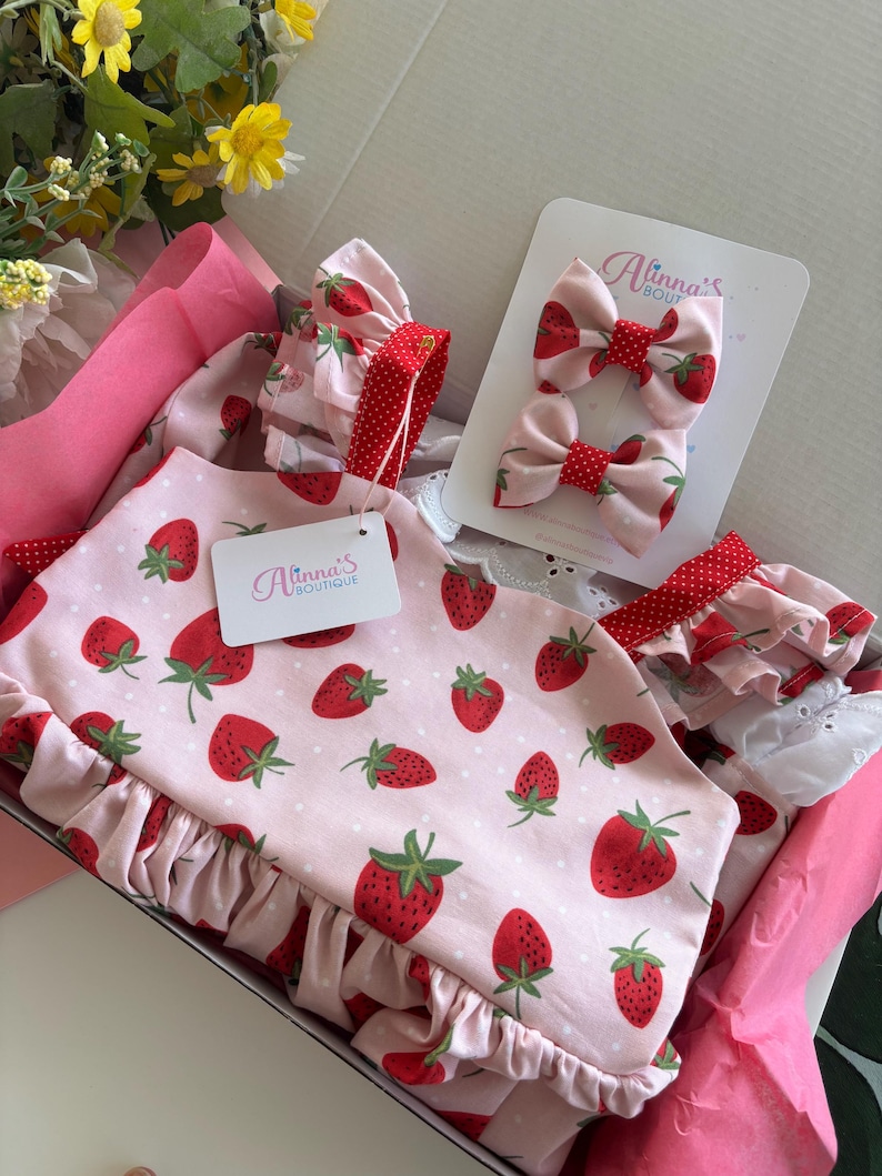 May include: A pink baby outfit set with a strawberry pattern, including a top, matching hair bows, and a ruffled trim. The set is presented in a pink box, with the brand name "Alinna's Boutique" visible on a tag.