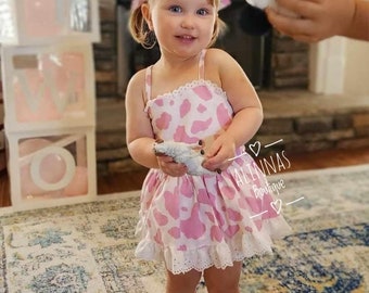 May include: A young girl in a pink and white cow-print dress and white fringed boots. The dress features a ruffled hem and eyelet lace trim. She is holding a toy. The background includes balloons and a sign that says "10".