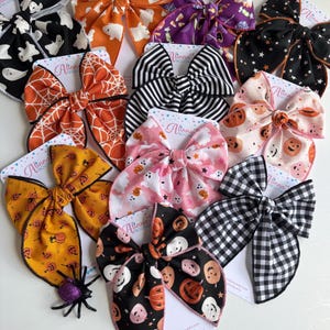 May include: A collection of Halloween-themed hair bows in various colors and patterns. Designs include ghosts, pumpkins, spiderwebs, and stars. The bows are made of fabric and are attached to cards with the brand name "Alinna Boutique".