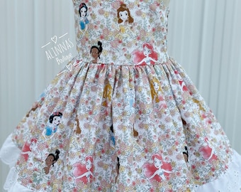 May include: A floral print dress featuring Disney princesses. The dress has a white base with a pattern of flowers and princess illustrations. It has ruffled sleeves and a white eyelet lace trim at the hem. The text "Alinnas Boutique" is visible.