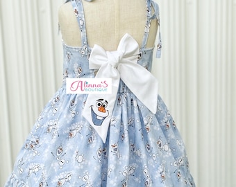 May include: A light blue sleeveless dress featuring an all-over print of Olaf the snowman and white snowflakes. The dress has tie-up shoulder straps, a large white bow at the back, and a ruffled hem. A logo reading Alinna's Boutique is visible.