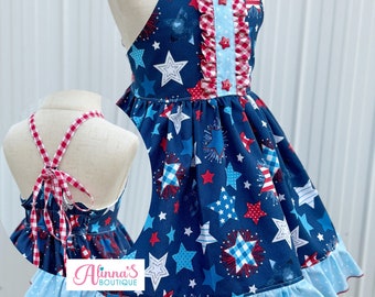 May include: A navy blue halter dress with a patriotic star and firework print. The dress features red and white gingham trim, a light blue polka dot ruffle, and red star-shaped buttons. The back ties with a gingham bow.