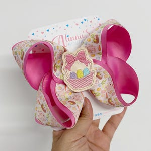 May include: A large, pink hair bow with a decorative Easter-themed patch. The bow features a patterned ribbon with bunnies and carrots, and a solid pink ribbon. The patch depicts a basket with Easter eggs. The bow is held in a person's hand.