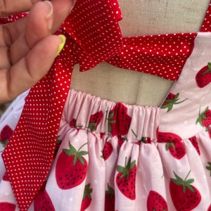 May include: A pink and red strawberry-themed dress. The dress features a pink fabric with a strawberry pattern and red polka dot bows. The dress has a gathered waist and tie straps.