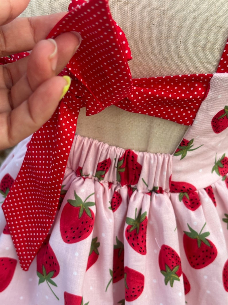 May include: A pink and red strawberry-themed dress. The dress features a pink fabric with a strawberry pattern and red polka dot bows. The dress has a gathered waist and tie straps.