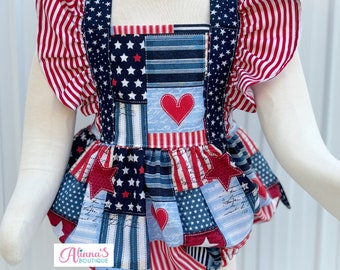 May include: A patriotic baby romper with a patchwork design featuring red, white, and blue stars and stripes. The romper has ruffled sleeves and a skirt, with a red heart applique on the bodice. The text "Alina's Boutique" is visible.