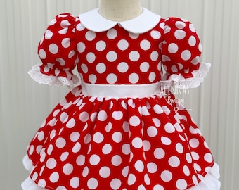 May include: A red and white polka dot dress with a white collar and short sleeves. The dress has a white ruffle at the bottom and a white sash around the waist. The dress is on a wooden mannequin.