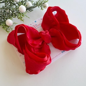 May include: A large, vibrant red hair bow made of soft fabric. The bow is layered and has a full, voluminous shape. It rests on a white card with the text "Alinn Boutique" and a website address. Decorative greenery with white ball ornaments is in the background.