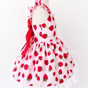 May include: A pink dress with a strawberry pattern. The dress features red strawberries with green leaves scattered across the fabric. It has a white lace trim at the hem and a red bow. The dress is displayed on a mannequin.