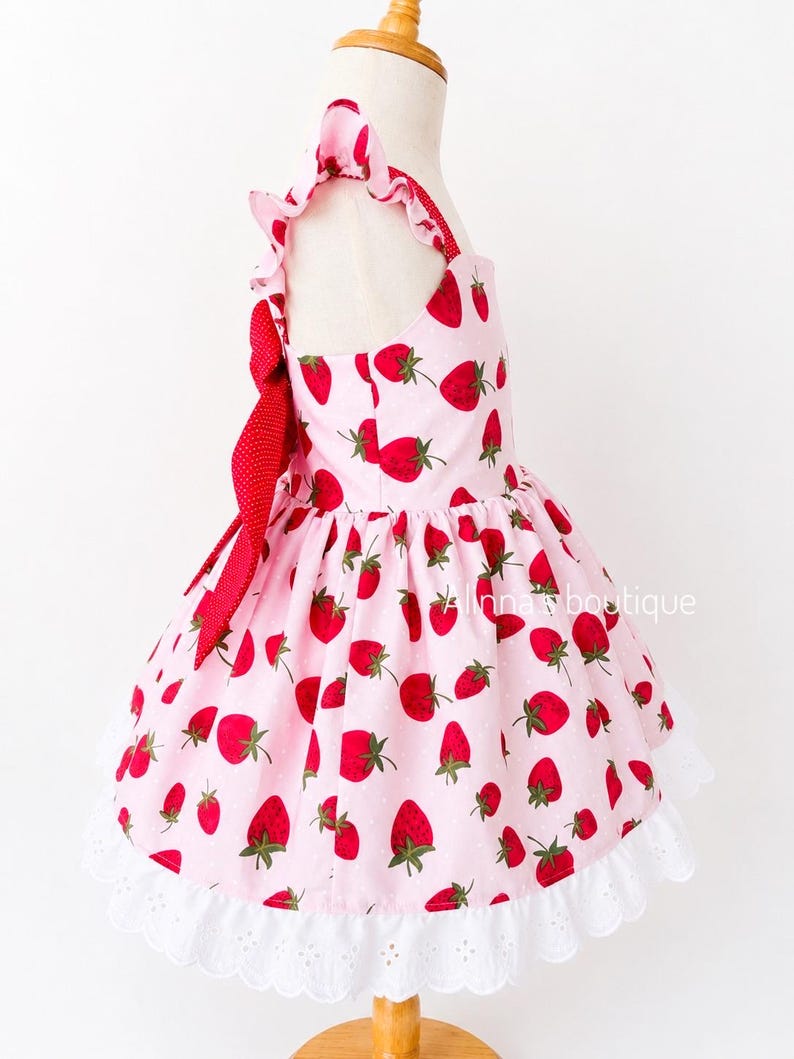 May include: A pink dress with a strawberry pattern. The dress features red strawberries with green leaves scattered across the fabric. It has a white lace trim at the hem and a red bow. The dress is displayed on a mannequin.