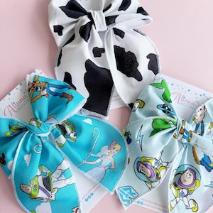 May include: Three fabric hair bows attached to branded display cards. One bow features a black and white cow print pattern. The other two bows display colorful cartoon character prints, including figures like a space ranger, a cowboy, and a shepherd.