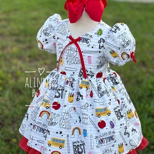 May include: White dress with a red bow and a school-themed print featuring yellow school buses, apples, pencils, and the words "Hey Hi Hello", "You're the Best", "Speak Up and Be Heard", and "I can do anything".