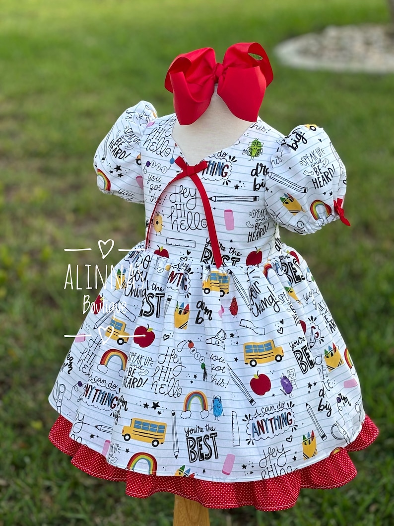 May include: White dress with a red bow and a school-themed print featuring yellow school buses, apples, pencils, and the words "Hey Hi Hello", "You're the Best", "Speak Up and Be Heard", and "I can do anything".