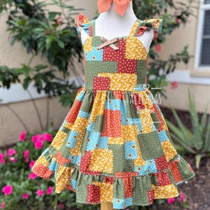 Puede incluir: Un vestido estilo patchwork con dobladillo con volantes y tirantes. El vestido presenta estampados florales y de lunares en rojo, amarillo, azul y verde. Un lazo color melocotón se asienta sobre el maniquí.