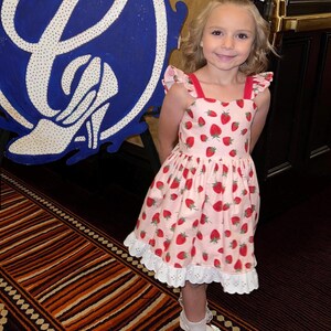 May include: A young girl wearing a pink dress with a strawberry pattern. The dress has red straps and white eyelet lace trim. She is also wearing a matching bow, white socks, and white shoes. A large blue and white sign is in the background.