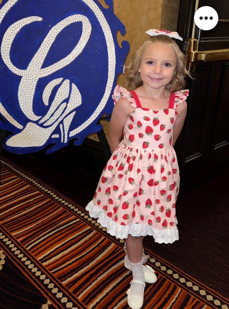 May include: A young girl wearing a pink dress with a strawberry pattern. The dress has red straps and white eyelet lace trim. She is also wearing a matching bow, white socks, and white shoes. A large blue and white sign is in the background.