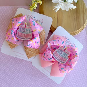 Puede incluir: Dos lazos para el pelo rosa y blanco con un diseño de cono de gofre y un parche de pastel "It's My Birthday". Los lazos están en tarjetas blancas con el nombre de la tienda, "Alinna's Boutique", impreso en ellas.