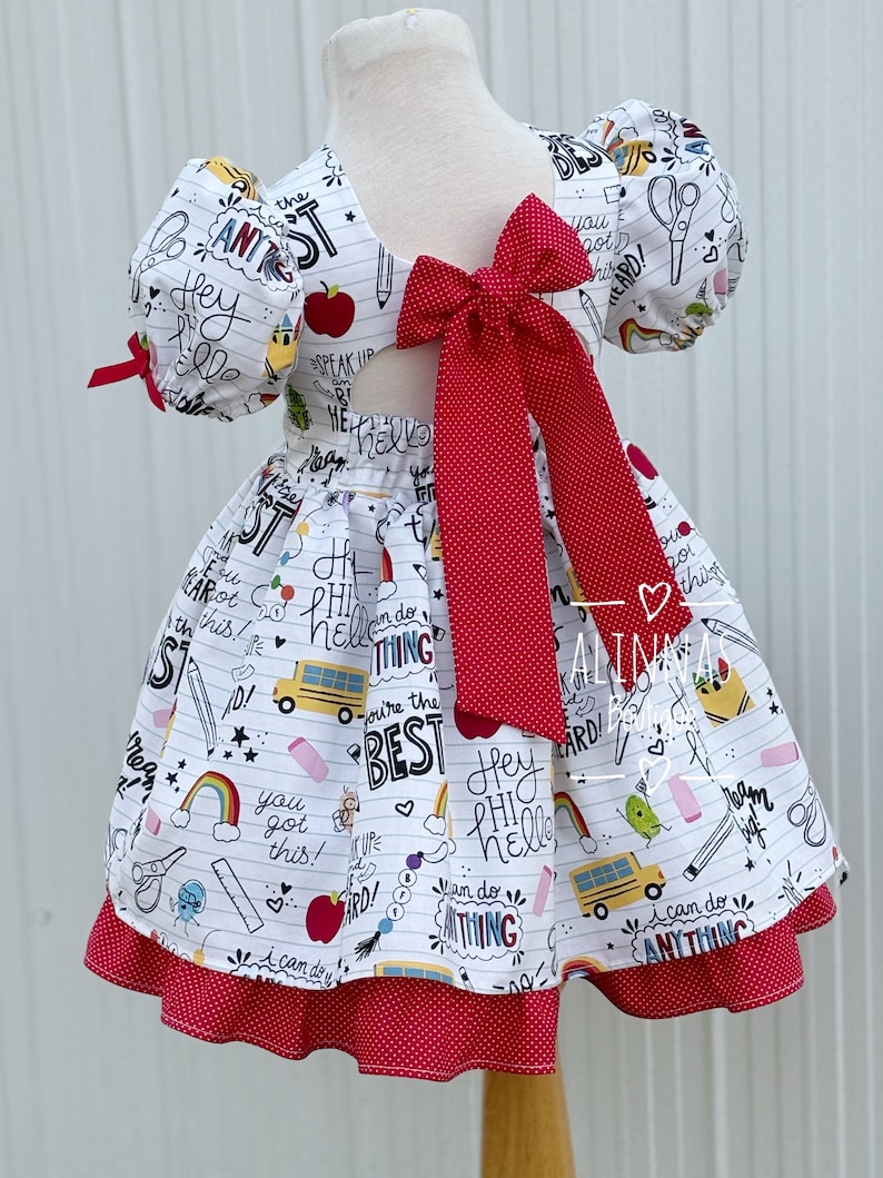 May include: A white dress with a red bow and a school-themed print. The print includes phrases like "Hey Phi Hello", "I can do anything", and "You're the best".