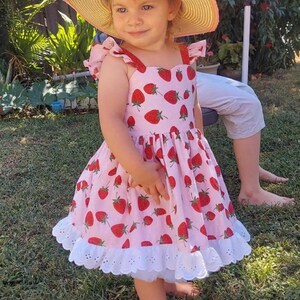 May include: A young child wearing a pink dress adorned with red strawberry patterns and a white eyelet hem. The dress has red straps and is paired with a straw hat. The child is standing on green grass.