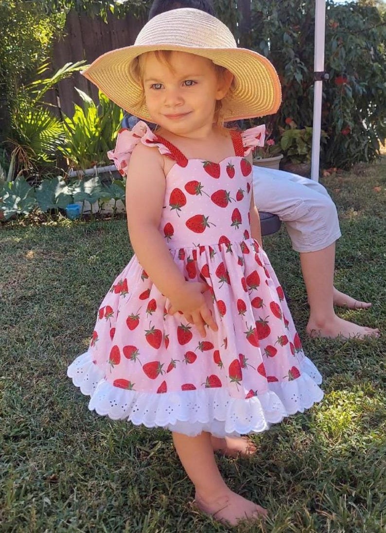 May include: A young child wearing a pink dress adorned with red strawberry patterns and a white eyelet hem. The dress has red straps and is paired with a straw hat. The child is standing on green grass.