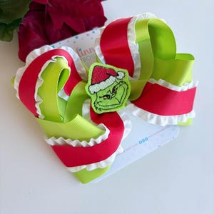 Peut inclure: Un nœud pour cheveux festif avec un motif du Grinch. Le nœud est composé de rubans rouges et vert lime superposés avec des bords blancs à volants. Le Grinch porte un bonnet de Père Noël. Le nœud est fixé à une carte blanche.