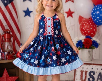 May include: A sleeveless dress featuring a navy blue fabric with a pattern of white, red, and blue stars, a central vertical ruffle with red star accents, and a light blue polka-dot ruffled hem. The dress has a halter-style neckline.