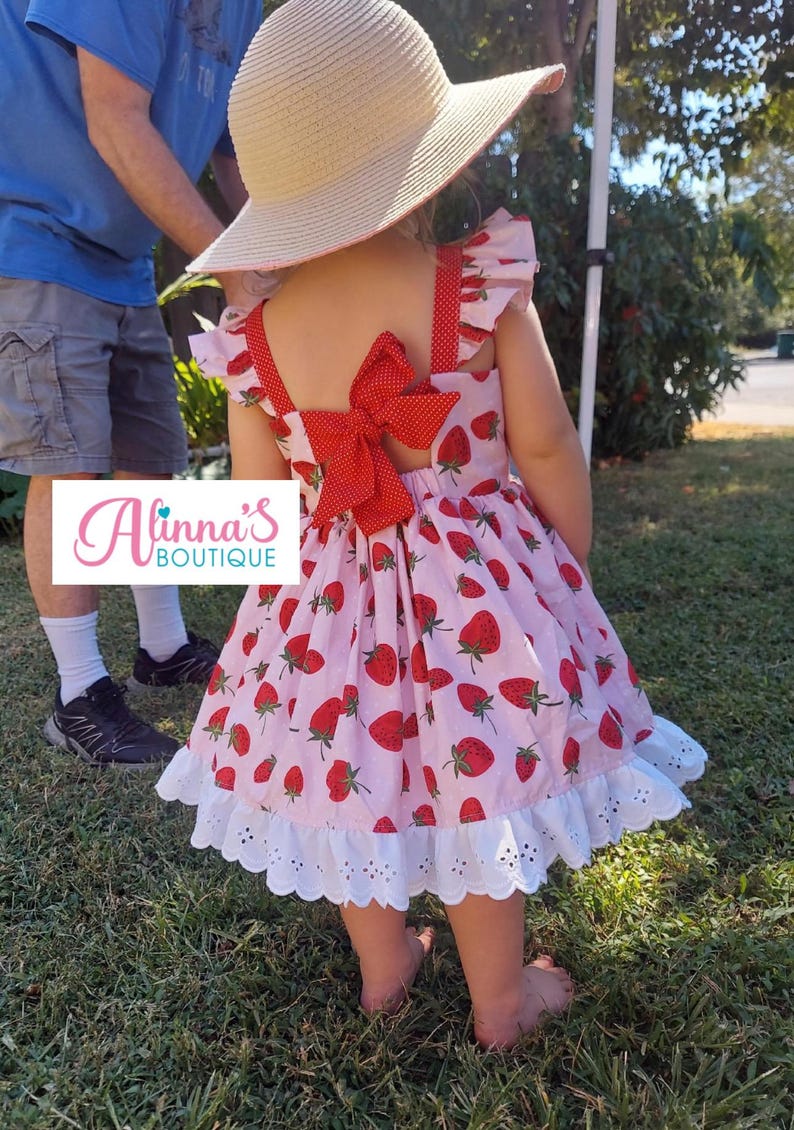 May include: A light pink dress with a strawberry pattern and a large red bow on the back. The dress has white eyelet lace trim and is worn with a straw hat. The dress is from Alinna's Boutique.