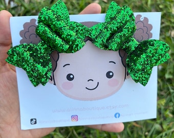 May include: Two green glitter hair bows with a cartoon illustration of a child's face on a white card. The card also includes the text "www.ainnaboutique.etsy.com" and social media icons for TikTok, Instagram, and Facebook.