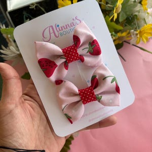 May include: Two pink fabric hair bows with a strawberry pattern and red polka dot centers. The bows are attached to a white card with the text "Alinna's Boutique". The background is pink and includes flowers.