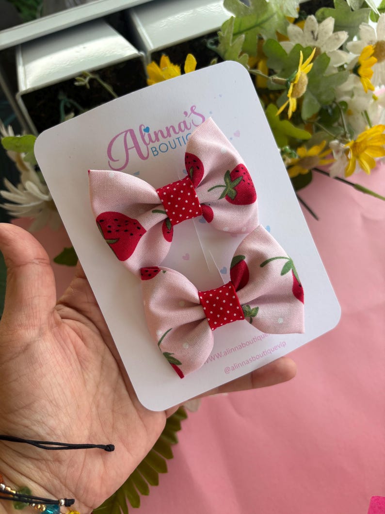 May include: Two pink fabric hair bows with a strawberry pattern and red polka dot centers. The bows are attached to a white card with the text "Alinna's Boutique". The background is pink and includes flowers.