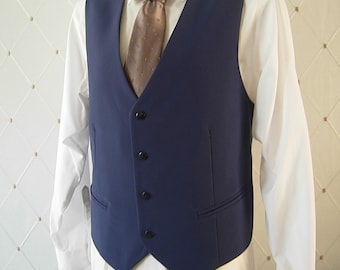 May include: A navy blue vest featuring a V-neckline and four black buttons down the front. The vest is worn over a crisp white long-sleeved collared shirt and is paired with a light brown necktie that has a subtle, repeating pattern.