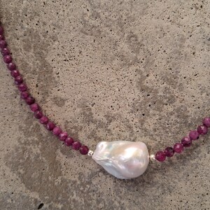 May include: A necklace featuring a large, irregularly shaped white pearl pendant. The necklace has a strand of small, round, deep pink beads. The beads are strung together with silver-colored metal. The background is a textured gray surface.
