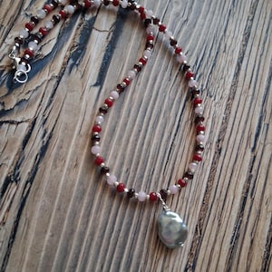 May include: A beaded necklace with a large, irregularly shaped, gray pearl pendant. The necklace features small, round beads in shades of red, pink, and silver, strung together. The clasp is silver. The necklace is displayed on a wooden surface.