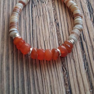 May include: A handmade bracelet featuring alternating sections of orange-red gemstone beads and cream-colored shell discs. The bracelet is strung on a flexible cord and accented with silver-colored metal rings. The beads and discs are approximately 0.25 inches in diameter.
